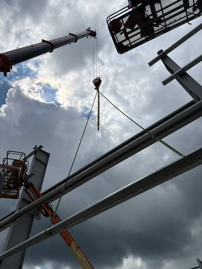 An aerial construction scene unfolds against a dramatic sky in Jodłownik, Poland. A large crane arm extends from the left, its hook suspended mid-air by chains and straps, ready to lift a heavy steel beam. Below, a cherry picker, painted orange, holds a worker wearing a yellow hard hat, assisting in the process. Multiple steel beams are visible, forming the framework of a building, with more in the process of being assembled. Dark, heavy clouds dominate the sky, interspersed with patches of bright sunlight, suggesting an impending storm. The overall atmosphere is one of active construction work in challenging weather conditions.