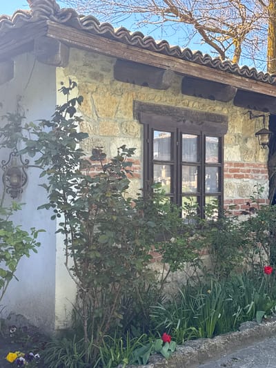The image shows the exterior of a building in Kalimantsi, Bulgaria. The building's facade is constructed of stone and brick. A wooden-framed window is visible on the wall. The roof is tiled and supported by wooden beams. Plants and flowers grow along the side of the building. A tree with bare branches is visible in the upper left corner of the image.