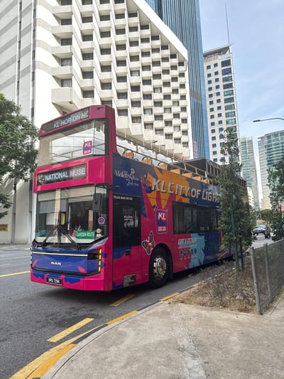 A pink and blue double-decker KL Hop On Hop Off city tour bus is parked on the right side of a street in Kuala Lumpur, Malaysia. The bus features advertisements and route information on its side, including "KL CITY OF LIGHTS." In the background, modern high-rise buildings dominate the skyline, including a white hotel with many balconies. The street is paved, with yellow and white lane markings visible. A sidewalk and a metal fence line the right side of the road. The sky is overcast. The license plate of the bus is VPQ 7294.