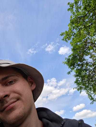 A selfie taken from a low angle shows a man's face, wearing a light brown hat, occupying the upper left portion of the frame. His gaze is directed slightly upwards. The right and lower portions of the image feature a bright blue sky with scattered white clouds and the green foliage of a tree. No specific location in Zhabinka, Belarus can be determined from the image.