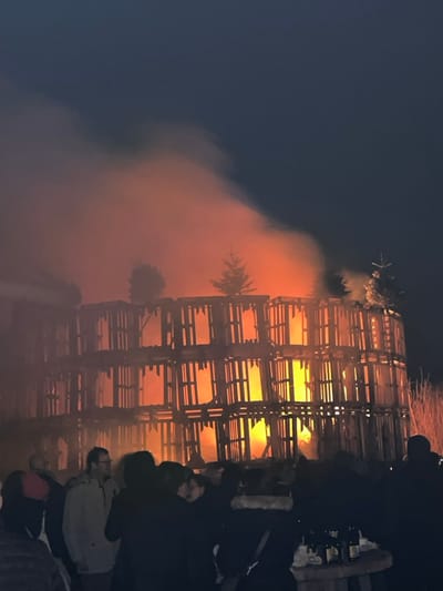 A night scene showcases a towering structure engulfed in flames. Constructed from a wooden grid, the fire illuminates the interior with an intense orange glow. Smoke billows around the blaze, partially obscuring the view. Silhouetted figures are gathered at the base of the structure, seemingly watching the spectacle unfold. The sky is a deep blue, contrasting with the fiery hues.