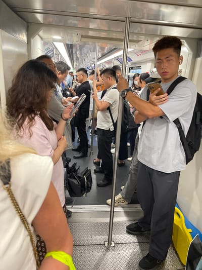 The image depicts a crowded subway car, likely during rush hour given the density of people. The subjects are a diverse group of individuals, primarily appearing to be of East Asian descent. They are dressed in casual attire, typical for commuting. Many are looking at their phones, suggesting they are passing the time during their journey. One person in the foreground is holding a smartphone and appears to be looking at it. Another person, also in the foreground, is seated and looking away from the camera, with a portion of their arm and leg visible. In the middle ground, several people are standing, holding onto overhead bars for support.
The setting is clearly an indoor public transportation vehicle, specifically a subway or train car, as indicated by the metallic walls, grab bars, and seating arrangements. The overall lighting is artificial and consistent with indoor lighting. There are no clear indicators of the time of day or weather conditions, though the crowded nature might suggest a peak commuting time. The emotions of the individuals are difficult to discern due to the angle and distance, but the general atmosphere appears to be one of routine transit.
Notable details include the various electronic devices people are using, suggesting a reliance on technology for entertainment or information. The presence of bags and backpacks indicates that people are traveling to or from work, school, or other destinations. A bright yellow and blue object, possibly a bag or luggage, is visible in the upper right corner, adding a splash of color to the otherwise utilitarian interior. There is no legible text visible in the image. The location context suggests this scene is taking place in the Longhua District of China.