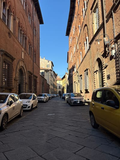 The image displays a narrow, stone-paved street in Lucca, Italy, bordered by multi-story buildings constructed primarily of red-brown brick. A clear blue sky is visible between the structures. The building on the left features multiple levels of arched windows with stone pilasters, its lower section partially in shadow. The building on the right exhibits similar architectural details, with strong sunlight illuminating its upper sections and casting prominent shadows. A decorative lamppost with a banner, displaying a crest and the word "MARTINI," is affixed to the right building. A bright yellow compact car is parked on the immediate right, partially out of frame. Numerous other compact cars of various light colors (white, silver, grey) are parked bumper-to-bumper along both sides of the street, receding into the distance. The street surface is composed of dark gray, rectangular stone blocks. A single, dark-clad individual is visible walking away in the far distance, positioned centrally between the parked vehicles.