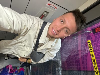 A selfie depicts a man inside what appears to be a passenger train or similar vehicle. He is wearing a light-colored jacket and has a dark-colored shoulder bag. Text in Russian, including the words "НЕ ПРИСЛОН" and "ТОРМОЗИТЬ," is visible on the train's interior. Based on the presence of Cyrillic script and context clues like passenger travel, the location is likely ДСК «Мичуринец», Russia.