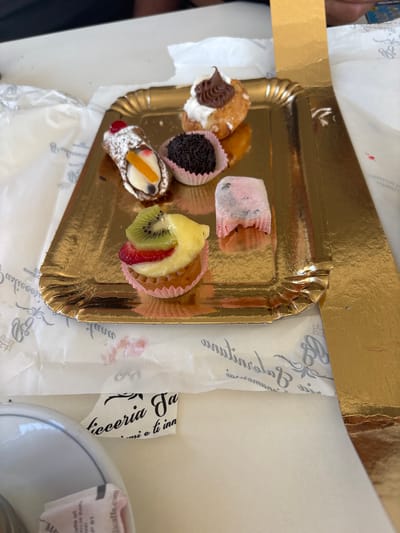 A high-angle shot shows a small assortment of pastries on a golden, fluted paper tray. The tray is placed on top of some white paper with decorative script writing. The pastries appear to be a variety of small, Italian-style treats.
From left to right, the pastries include: a cannoli filled with white cream and topped with chocolate icing and sprinkles, another cannoli topped with a cherry and orange slice, a small pastry with a pink paper liner and dark chocolate sprinkles, a pink and white swirled pastry, and a mini tart topped with kiwi, strawberry, and what looks like pineapple.
The scene appears to be an outdoor setting, possibly a cafe or bakery in Ardea, Italy, given the context. The lighting suggests it is daytime, though no specific weather conditions are evident. The overall impression is of a delightful treat selection, perhaps a light snack or dessert.
Visible text on the white paper includes "Pasticceria Ga" and "mi e ti inn," suggesting the name of the establishment where the pastries were purchased. There is also some text on a small white packet to the right, which is partially obscured but appears to be related to coffee.