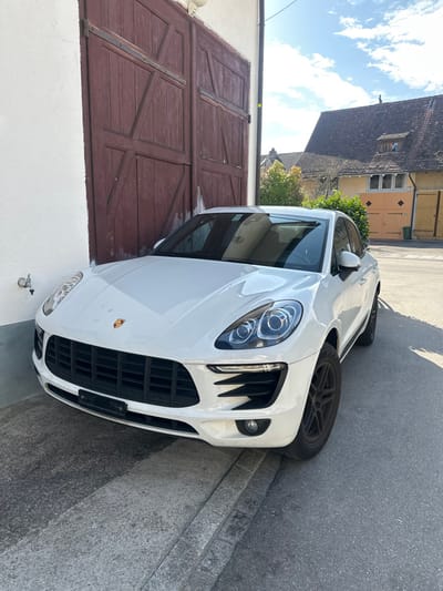 The image displays a white Porsche Macan SUV parked in Rafz, Switzerland, under a bright daytime sky. The vehicle is positioned with its front towards the viewer and slightly to the left, resting partially on a concrete ramp and an adjacent asphalt road, with its front wheels on the asphalt. The SUV features a visible Porsche crest on its hood and dark-colored multi-spoke wheels. To the immediate left of the car's front section, a silver-toned water spigot is mounted on a white building wall. Directly behind the vehicle are large, weathered reddish-brown wooden double doors, integrated into the white building structure. In the background, under a blue sky with scattered white clouds, various structures are visible. A prominent background building features a dark tiled pitched roof with dormer windows, a light yellow facade, and a set of lighter wooden double doors at ground level. A dark, upright object is discernible on the peak of this background building's roof. Green foliage from trees and shrubs is interspersed among the distant structures. No individuals are present in the scene.