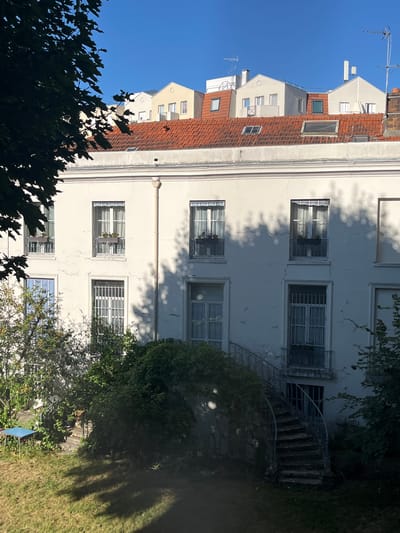 This image captures a view of the rear facade of a building in L'Haÿ-les-Roses, France, seen from a garden. The building is painted a light cream color and features multiple windows with white frames. Some windows on the upper floor have decorative flower boxes with red flowers. The lower windows are secured with metal bars. A winding exterior staircase with a metal railing leads up to a doorway on the right side of the building. The building is partially obscured by lush green foliage and tree branches in the foreground, with a patch of grass and a small blue table visible in the garden. The roof of the building is covered with red tiles, and beyond it, several other buildings with different rooflines and dormer windows are visible against a clear blue sky. The overall impression is that of a quiet, residential area bathed in sunlight, with shadows cast by the trees and foliage on the building and lawn.