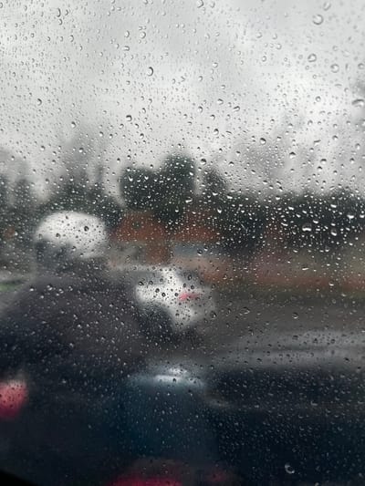 The image depicts a rainy street scene in Bondy, France, viewed through a transparent surface heavily covered in water droplets. The foreground is sharply rendered with numerous spherical and irregularly shaped raindrops of varying sizes. The background is significantly blurred due to both the wet viewing surface and depth of field. Visible in the blurred mid-ground are multiple vehicles on a dark, wet road. A light-colored car is present, featuring a discernible red taillight on its rear-right side. To the left of this car, a heavily blurred figure, possibly a person on a motorcycle or scooter, is suggested by a light-colored helmet-like shape. Further in the background, indistinct dark green foliage and reddish-brown architectural structures are visible under a uniformly grey, overcast sky. No specific human actions or interactions are clearly discernible. The lighting is diffuse, indicative of an overcast, rainy day.