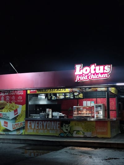 This nighttime image captures an illuminated "Lotus fried chicken" food stall in Leuwiliang, Indonesia, operating under a dark sky. The prominent red and white neon sign for "Lotus fried chicken" glows brightly above the stall. The structure features a dark red upper fascia and a lower counter area bustling with activity.
The counter displays various menu items on digital screens and physical boards, showcasing fried chicken, burgers, and fries. Inside the stall, kitchen equipment including a large exhaust hood, prep surfaces, and what appears to be warming display cases for cooked food are visible. On the left side, a large poster advertises a burger and french fries, with text that includes "HALALAN THAYYIBAN," indicating food prepared according to Islamic dietary laws. A phone number, 0857-7337-6276, is also visible on the stall's facade.
The front of the counter features a vibrant banner with the slogan "EVERYONE LOVE IT LIKE IT WANT IT" alongside a cheerful cartoon character of a boy next to a burger. The ground in front of the stall is asphalt, and a large puddle reflects the stall's lights, suggesting recent rain. No people are visible in the image, but the well-lit and fully equipped stall implies it is open for business. The scene conveys a lively, accessible fast-food establishment ready to serve customers.