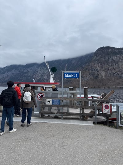 An outdoor scene depicts a ferry terminal identified by a prominent blue sign reading "Markt 1" in Hallstatt, Austria, situated on the Hallstättersee lake. The sky is overcast, with low-hanging clouds obscuring the upper portions of the steep, forested mountains in the background.

A red and white passenger ferry boat is docked at a wooden pier with metal railings. A white pole structure, possibly containing a navigation light or sensor, stands on the pier. Several informational and prohibitory signs are affixed to the pier railings: a red circular "no entry" symbol, a white square with a black dog-on-leash icon, and a red circular sign stating "Parken verboten!" (No parking!).

Six individuals are visible. Two persons are in the foreground, standing on a grey paved surface facing the pier from behind. One wears a dark jacket, blue jeans, and a black backpack. The other wears a brown jacket, blue jeans, and a white backpack. Four other individuals are near or on the ferry boat. One person in a red jacket stands on the pier near the ferry's entrance. Another individual is partially visible with a yellow hooded garment. A person with dark hair is seen inside the ferry's cabin, and another figure is partially obscured behind the pier's railing. The individuals appear to be passengers waiting or boarding the ferry.