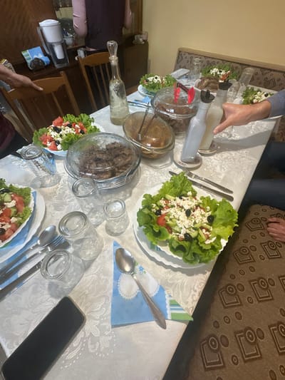 This image captures an indoor dining scene in Letnitsa, Bulgaria, featuring a table laden with food and beverages. The main subject is a rectangular dining table covered with a white tablecloth featuring an embossed floral pattern.
**People:**
At least three individuals are partially visible. On the left, a person's arm in a dark red/purple garment rests near a wooden chair. On the right, a person's hand and forearm, clad in a light blue long-sleeved shirt, reach towards two white plastic bottles. In the upper background, the back of a person wearing a pink top and dark vest is visible next to a dark wooden cabinet. On the far right, a person's feet and ankles are seen resting on a brown patterned sofa or bench.
**Food and Beverages:**
The table holds multiple dishes and drinks. Several plates of salad are prominent, consisting of green lettuce, diced tomatoes, sliced cucumbers, black olives, and crumbled white feta cheese. Two large clear glass bowls with domed lids contain cooked brown meat, possibly meatballs. Another clear glass bowl, uncovered, holds a brown-colored dish with a spoon. A tall, clear glass decanter with an ornate stopper contains a clear liquid. Two white opaque plastic bottles with dark pour spouts are present, one being reached for. Numerous empty clear glass drinking tumblers are scattered across the table. In the background, on a dark wooden cabinet, a white pitcher-style water filter is visible.
**Objects and Environment:**
The table is set with silverware, including forks, knives, and spoons, placed on blue and white patterned cloth napkins. A black smartphone with a metallic edge rests face down on the tablecloth in the lower left foreground. The background features a dark wooden cabinet/dresser against a light yellow wall. To the right of the table, a brown sofa or bench with a repetitive geometric pattern is visible. The lighting is consistent with an indoor setting.