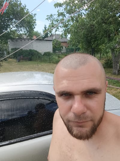 A man with a shaved head and a beard is taking a selfie. He is shirtless and appears to be outdoors. In the background, there is a white brick building with a red chimney, some trees, and a fence. A silver car is visible in the foreground, with soap suds on it, suggesting the man may be washing his car. The sky is blue with some clouds, indicating it is daytime and likely fair weather. The man's expression is neutral. There is no visible text in the image. The scene appears to be in a rural or suburban setting in Pishchane, Ukraine.