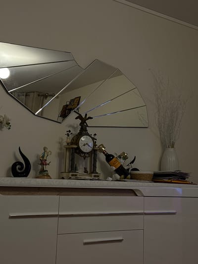 The image displays an interior residential scene in Colombes, France. A light-colored wall forms the background, against which a white, modern-style cabinet with multiple drawers and rectangular silver handles is positioned. Above the cabinet, a large, multi-segmented mirror with a fan-like geometric design is mounted, reflecting interior elements such as curtains and framed artwork.
The top surface of the cabinet serves as a display area for various decorative objects. From left to right, these include: a partially visible white floral arrangement; a black, abstract spiral-shaped sculpture; a colorful ceramic figurine depicting a young boy in historical clothing holding an object, standing on a pedestal; and an ornate antique mantel clock, featuring columns, a visible clock face, and a golden eagle figure atop its structure. To the right of the clock, a white figurine is shown holding a dark-colored wine bottle, identifiable as Veuve Clicquot Champagne by its orange label. Adjacent to this, a small woven basket and a stack of papers or magazines are present. The far right side features a tall, white fluted vase containing numerous white decorative branches. The scene is lit from the left, creating shadows and highlights on the objects. No people are depicted.