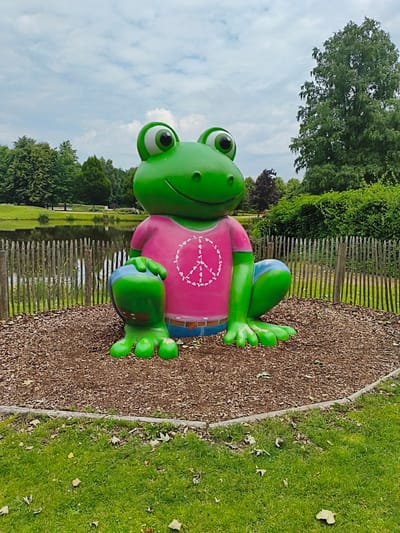 The image shows a large, brightly colored statue of a frog wearing a pink t-shirt with a white peace symbol and blue shorts. The frog is positioned outdoors, in what appears to be a park or garden setting. It is sitting on a circular area covered with wood chips, which is bordered by a wooden fence. Behind the fence, there is a body of water, likely a pond or canal, and a grassy area with trees and bushes. The sky is cloudy. The statue is the central and dominant subject of the image. Based on the context of the city of Papenburg, Germany, this statue is located in the popular tourist attraction "Meyer Werft", specifically within the visitor center's outdoor area.