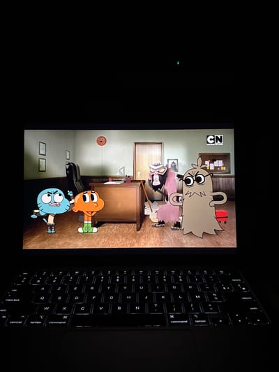 The image shows a laptop screen displaying an episode of "The Amazing World of Gumball." The scene appears to be set inside Principal Brown's office, with Gumball and Darwin standing near his desk. Principal Brown and Ms. Simian are also in the scene. A keyboard is visible to the right of the screen. The laptop is likely located indoors, with the room dimly lit. No location information for Champigny-sur-Marne, France is available from the image.