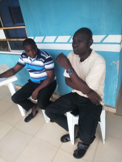 Two dark-skinned adult males are seated on white plastic stools in front of a blue and white striped wall. The man on the left wears a blue, white, and black striped polo shirt and black pants. He is seated at a white table, with his left arm resting on it. He is looking down at the table. The man on the right wears a tan t-shirt and black pants. He is looking to his right, with his arms crossed. A watch is visible on his left wrist. The floor is tiled with light beige squares. A dark-colored flip phone is visible on the seat of the stool of the man on the right. The overall environment appears to be outdoors, possibly in a public space like a waiting area. The context of Ajasse Ipo, Nigeria, implies this could be a governmental or administrative building, or a commercial establishment within the town.