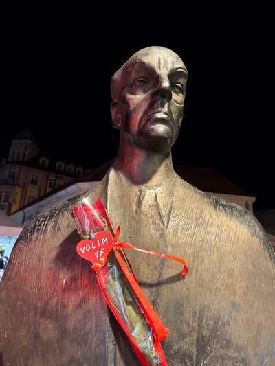 The image shows a close-up, low-angle, night-time shot of a bronze bust of a man. A single red rose, wrapped in clear plastic and tied with a red ribbon, is affixed to the chest of the bust. A small red heart-shaped tag with the words "VOLIM TE" written in white is attached to the ribbon. The bust is textured and shows signs of age and wear. In the background, slightly out of focus, are the architectural details of buildings, suggesting an urban setting. The lighting is dramatic, with red and purple hues cast on the bust from an unseen light source, contrasting with the dark night sky. The overall impression is solemn and romantic. This bust is located in Skver, a public square in the city of Tuzla, Bosnia and Herzegovina, and is a tribute to Meša Selimović, a renowned Yugoslav writer.
