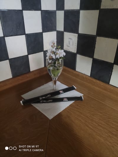 This is a forensic, matter-of-fact description of the image, which depicts a still life composition on a wooden countertop. The background features a black and white checkered tile wall, typical of a kitchen or food establishment. In the center of the frame, a clear glass contains a small arrangement of white flowers with green stems and buds. Two black, individually wrapped chopsticks with red Chinese characters are placed on a white paper napkin below the glass. The wooden surface has a warm, reddish-brown tone with a visible grain pattern. The lower left corner of the image shows the text "SHOT ON MI 9T AI TRIPLE CAMERA," indicating the device used to capture the photograph. There are no people or actions depicted in the scene. The overall impression is one of a tidy and perhaps decorative setting, suggesting a close-up view of items intended for dining. The location within Bagnoli di Sopra, Italy, is not discernible from the visual elements of the image itself.