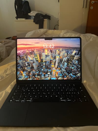 The image displays an Apple iPad, encased in a black keyboard accessory (likely a Magic Keyboard), resting on a rumpled white bedsheet. The scene is set indoors, likely a bedroom, in Basiglio, Italy. The tablet's screen is active, showing a lock screen with a dynamic wallpaper of a brightly lit city skyline, possibly at dusk or night.

Prominently displayed on the screen is the date "Jeudi 26 décembre" (Thursday, December 26) and the time "13:42" (1:42 PM). The tablet's status bar shows a battery icon indicating a charge level around 80%, along with partially visible text "Suivre rond." The keyboard features a French layout, with various keys illuminated, including "control," "option," and "command."

In the background, to the left of the tablet, a light-colored wardrobe or closet door is visible, alongside a pile of dark clothing. The tablet is in an idle state, not being actively operated. The overall lighting suggests an indoor setting with artificial light or dim natural light.