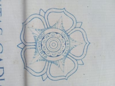 A blue, circular, stylized floral or mandala pattern is imprinted on a white, woven textile. The pattern features an outer perimeter of eight connected, rounded petal shapes, an inner eight-pointed star motif, and multiple concentric circles with dashed and solid line details culminating in a small central circle. The blue coloration, exhibiting minor variations in density, appears to be composed of short dashed segments, consistent with a stamp or print. The underlying fabric is off-white, displaying a prominent, uniform grid-like weave texture. Sparse dark particulate matter is scattered across the textile surface. This image depicts an object located in Teupinjok, Indonesia.