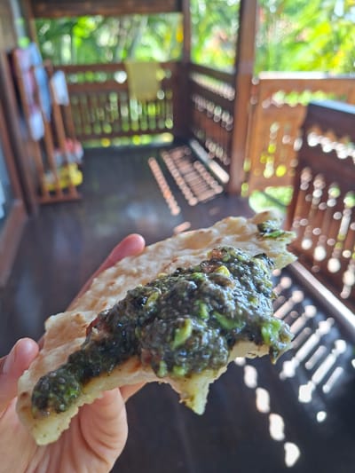 A person's hand holds a piece of flatbread topped with a dark green, textured spread. The location is Lodtunduh, Indonesia, suggested by the wooden structure with sunlight filtering through it in the background, a common architectural element in the region. The structure is open-air, possibly a gazebo or veranda. In the out-of-focus background, there's greenery visible. The wooden structure and surrounding foliage point to a natural or semi-rural setting. The focal point is the food, which suggests the activity is either eating or food preparation and presentation.
