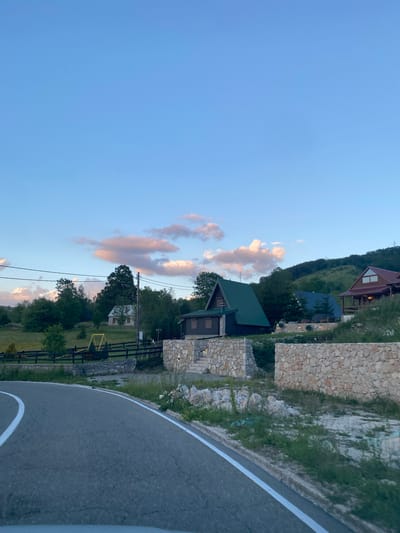The image depicts a rural mountain landscape near the city of Praga, Montenegro, during what appears to be late afternoon or early evening. A paved road with white lane markings curves through the foreground, suggesting a viewpoint from a vehicle. Beyond the road, a natural landscape unfolds, featuring rolling hills covered in lush green trees. Several wooden houses with distinct architectural styles are scattered across the hillside, some with steep, dark roofs and others with lighter-colored wood exteriors.

One prominent house in the mid-ground features a dark green, pitched roof and appears to be constructed from dark wood logs. Adjacent to this, a smaller, lighter-colored house is visible among the trees. Further up the hill, another wooden house with a prominent red roof and large windows is situated, appearing to be a more substantial dwelling. The houses are interspersed with natural elements, including trees and bushes, and some are bordered by low, dry-stone walls made of light-colored, irregular stones.

In the background, the sky is a clear blue with scattered clouds tinged with pink and orange, indicating the setting sun. The overall atmosphere is serene and tranquil, characteristic of a mountainous rural setting. There are no people or animals visible in the image. The environment is clean and well-maintained, with a focus on natural materials and integration with the surrounding environment.