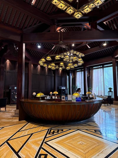 The image depicts an upscale interior identified as a reception area or lobby within a building in Tongxiang, China.

A prominent semi-circular reception desk, crafted from dark polished wood with a vertically fluted base, occupies the midground. Its top surface displays three illuminated Tiffany-style lamps with amber-colored glass shades, multiple informational signs/stands, a small bell, and a plant. Behind this desk, one individual, with short dark hair and wearing a light-colored shirt, is visible standing, positioned towards the right side of the frame.

Overhead, a large, multi-tiered chandelier with numerous illuminated amber-colored, faceted glass shades is suspended. Additional square-shaped chandeliers with similar glass elements are integrated into the dark wooden ceiling, which features exposed beams and slatted panels. Recessed spotlights are also visible in the ceiling. The walls are clad in dark wooden paneling with vertical accent lighting. To the right, tall windows are covered by light-colored, translucent drapes, admitting diffused natural light. The floor consists of highly polished marble or stone tiles in cream and amber tones, distinguished by dark geometric patterns.

Further objects include a dark armchair and a wooden display cabinet on the left. On the right, beyond the reception desk, a cushioned seating area and a potted plant on a stand are partially visible.