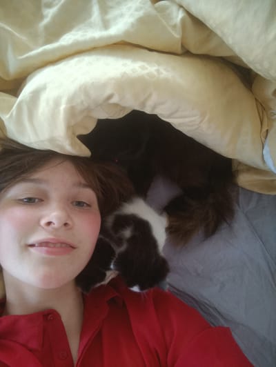 A young person with brown hair and light skin is lying down and looking at the camera, smiling. They are wearing a red shirt. Beside them, partially obscured by blankets, is a dark brown and white cat. The cat's fur is visible, as are its paws. They are lying on a bed with light yellow and gray bedding. The image appears to be taken indoors during the daytime. The atmosphere is relaxed and cozy, suggesting a moment of quiet comfort between the person and their pet. There is no visible text in the image.