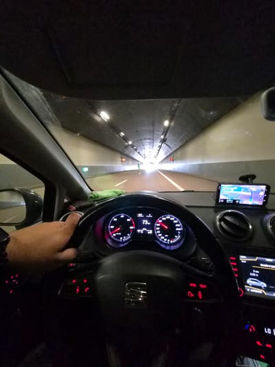 The image shows a first-person perspective of someone driving a car through a brightly lit tunnel. The car's dashboard is visible, displaying the speedometer indicating a speed of 73 km/h. A navigation system is mounted on the dashboard, showing a map with a route highlighted. The driver's left hand is on the steering wheel. The tunnel interior is characterized by a series of lights along the ceiling, illuminating the road ahead. The location is identified as being in Mondorf-les-Bains, Luxembourg.