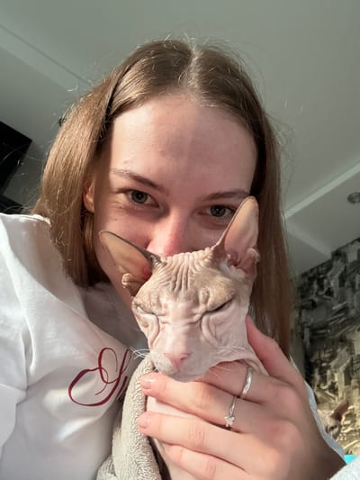 A young woman with long brown hair is holding a light-skinned Sphynx cat. The cat's eyes are closed, and its head is resting on the woman's chest, partially obscuring her face. The woman is wearing a white hooded sweatshirt with a burgundy decorative script. She has two silver rings on her right hand. The background shows a wall with patterned wallpaper and a ceiling. The scene appears to be indoors. The image was taken in Novaya Ivanovka, Russia.