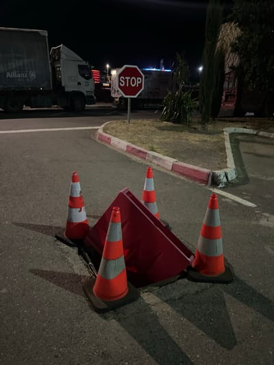 The image shows a street scene at night in Jbilate, Morocco. A red, rectangular object, potentially a container or barrier, is surrounded by four traffic cones with alternating orange and white stripes. A 'STOP' sign is visible in the background, next to a large truck with 'Allianz' branding on its trailer. The street is paved, with a curb and a grassy area with trees and shrubs adjacent to it. The lighting is dim, provided by streetlights and the truck's headlights.