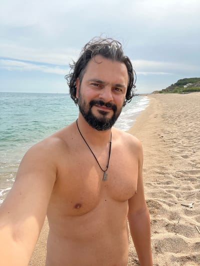 A man with dark, wavy hair and a beard stands on a sandy beach in Sant Pol de Mar, Spain. He is shirtless, wearing a necklace with a rectangular pendant. The man is looking at the camera and smiling. The ocean is to his left, with gentle waves rolling onto the shore. The beach is wide and sandy, with a few pieces of driftwood scattered about. In the background, there is a gentle slope covered in trees and some white buildings. The sky is cloudy.