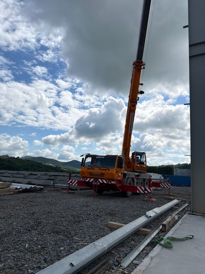 In Wilkowisko, Poland, a large orange Grove crane is positioned on a gravel construction site. The crane's boom is extended upwards, reaching towards a cloudy sky with patches of blue. In the background, lush green hills and trees form a natural landscape. The foreground of the image shows stacks of metal beams and wooden planks, suggesting ongoing construction work. A portion of a gray building is visible on the right side of the frame. The scene appears to be a construction site in a semi-rural or suburban area, with the natural environment contrasting with the industrial equipment. There are no people visible in the image.
