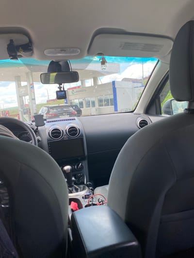 The image captures the interior of a car, looking forward through the windshield. A gas station is visible outside, with pumps and a building. A black Nissan Qashqai with the license plate "H647HY 702" is parked at one of the pumps. Inside the car, a dashcam is mounted on the windshield, and a rearview mirror is visible. The dashboard displays various controls and a screen. The seats in the foreground appear to be dark gray. The scene suggests a stop at a gas station, possibly in or near Ishmurzino, Russia, based on the context provided.