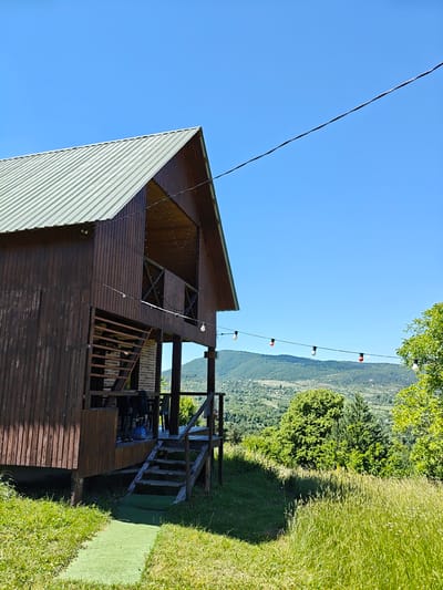 This media file depicts a wooden cabin situated on a lush, green hillside with a panoramic view of rolling, verdant hills under a clear blue sky. The cabin has a prominent gabled roof and is made of dark-stained wood. A wooden staircase leads up to a balcony area. String lights with colored bulbs are strung across the scene, suggesting an evening ambiance, though the bright sky indicates it is daytime. The foreground is dominated by tall, wild grasses, with a narrow strip of artificial turf leading towards the cabin's entrance. The background features a hazy, mountainous landscape dotted with small buildings and trees, indicative of a rural or semi-rural setting. The overall impression is one of a peaceful, natural retreat. No people or text are visible in the image.
