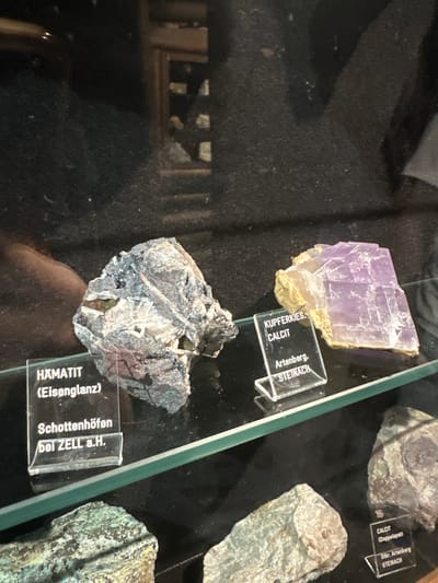This image captures a display of minerals, likely in a museum or educational setting, featuring specimens of Hematite, Calcite, and Kupferkies. The focus is on two prominent display cases containing the minerals. In the foreground, a rough, metallic-looking specimen labeled "HAMATIT (Eisenglanz)" rests on a shelf, accompanied by a label indicating its origin as "Schottenhöfen bei ZELL a.H." To its right, a cluster of translucent, lavender-colored crystals, identified as "KUPFERKIES, CALCIT" with the origin "Artenberg, STEINACH", is also displayed. Below these, other mineral samples are visible, one with a label indicating "CALCIT (Doppelspat) from Stbr. Artenberg STEINACH." The environment appears to be dimly lit, with the specimens displayed on glass shelves, suggesting a formal exhibition. The background is dark and out of focus, hinting at the broader context of the display area. The specific location is identified as the Mineralienmuseum in Schonach im Schwarzwald, Germany, further contextualizing the exhibit.
