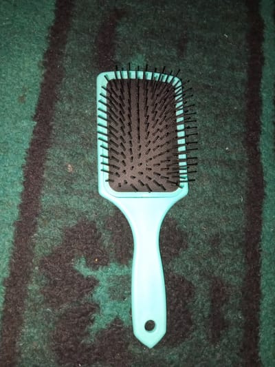 A light blue paddle brush with black bristles lies flat on a dark green and black striped rug. The brush is oriented vertically, with the handle at the bottom and the head at the top. The bristles are arranged in neat rows and appear to be made of plastic. The rug has a shaggy texture and the stripes are uneven in width. The scene appears to be indoors, possibly in a home, and there are no people visible. The lighting suggests it is daytime. The image does not contain any information that places it in Erzin, Turkey.