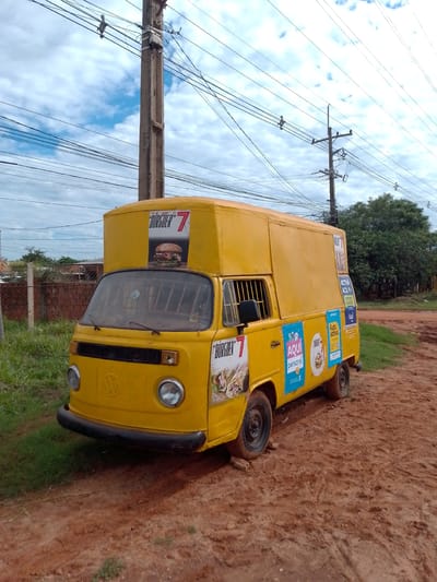 The image shows a yellow Volkswagen van, heavily modified with an expanded upper section, parked on a red dirt road. The van is adorned with advertisements, including one for "Burguer 7" featuring a hamburger image. Other ads include the "personal" logo and "Activa" text. The van appears to be in a state of disrepair, with dirty tires. The location is likely in Concepción, Paraguay, based on the presence of Spanish-language advertisements common in that area.

In the background, a telephone pole is visible with numerous wires. Patches of green grass border the road and a rudimentary fence can be seen in the mid-ground. Trees are visible further away. The sky is overcast with a mix of white and gray clouds. The image appears to have been taken during the daytime. No people are visible in the scene.