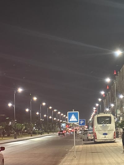 The image captures a nighttime street scene in Berrechid, Morocco. A white bus is parked on the right side of the road, facing away from the camera. Adjacent to the bus, a blue parking sign is visible. In front of the parking sign, a pedestrian crossing sign is displayed. The street is lined with trees and streetlights, casting a warm glow on the pavement. Several cars are on the road, their headlights illuminated. The sky above is dark. The overall impression is that of a typical urban environment at night.