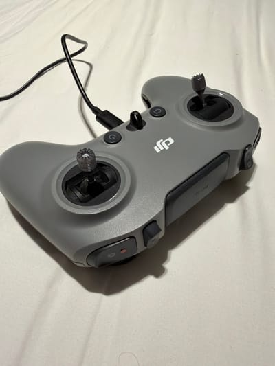 The image displays a gray DJI remote controller on a white, textured surface, likely a bedspread. The controller features two joysticks, several buttons, and a USB-C port with a black cable connected. The DJI logo is visible on the front of the controller. The lighting suggests an indoor setting, with a shadow cast by the controller. No people are present in the image. The location in Ban Phonsavang Neua, Laos, is not discernible from the visual information provided in the image.
