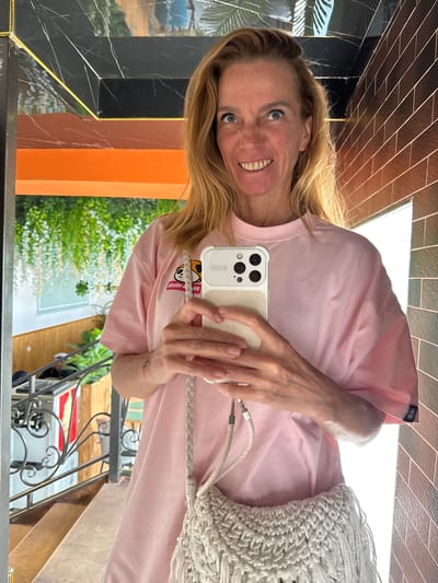 The image is a mirror selfie featuring a smiling woman with long blonde hair and blue eyes. She is wearing a light pink t-shirt with a small dark patch containing a white logo on her upper left arm. In her hands, she holds a light-colored smartphone with a distinctive triple camera setup on the back, attached to a beige woven strap. A large, off-white macrame bag is draped over her right shoulder. A silver ring is visible on her right hand. Her expression is joyful, with crinkled eyes reflecting a genuine smile.

The mirror's reflection showcases a diverse setting. On the left, a black tiled wall with white veining and gold trim is visible, alongside an orange structural element. Above this, dark brown or black rectangular tiles create a patterned surface. Beyond these architectural features, lush green foliage and bright white light are reflected, suggesting an outdoor garden or a significant window. On the right side of the reflection, ornate black metal railings are present, hinting at a balcony or staircase, and a red and white object is partially visible below. The phone's case prominently displays reversed text, "PP HOUSE," beneath a cartoon character. The overall bright illumination indicates a daytime setting. The scene captures a casual, happy moment within Semenovka, Kyrgyzstan.