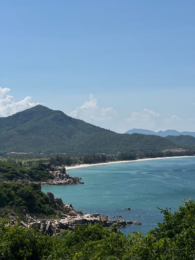 A panoramic, high-angle view captures a serene coastal landscape under a bright blue sky with scattered white clouds. The foreground is dominated by lush green foliage and rough, grey rock formations, creating a natural framing for the scene. Beyond this, a crescent-shaped beach with fine, pale sand stretches out, meeting the calm, turquoise waters of the sea. In the middle ground, a range of verdant, tree-covered hills rises, with a larger, more prominent mountain forming the backdrop. Along the coastline, a line of pine trees separates the beach from the foothills. Hints of buildings, possibly resorts or housing, can be seen nestled amongst the trees on the lower slopes of the hills. In the distance, a series of layered, blue-hued mountains fades into the hazy horizon. The image appears to be taken from an elevated position looking out over the sea towards the mountains, likely in the Vĩnh Hải area of Vietnam, known for its beautiful coastline and hilly terrain. There are no people visible in the image.