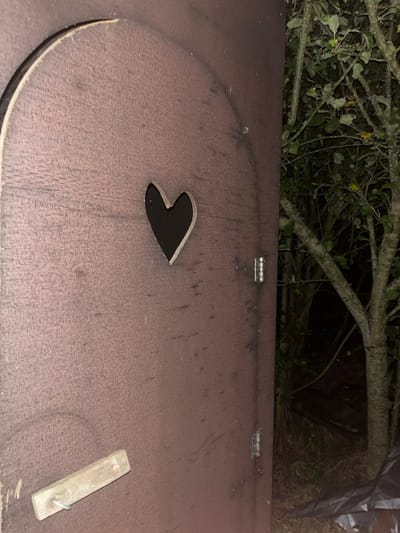 The image depicts a close-up view of a reddish-brown wooden door in a dimly lit outdoor setting, located in Sint-Pieters-Leeuw, Belgium. The door, which appears to be constructed from plywood or a similar composite, features an arched top edge. A distinct heart-shaped cutout, revealing the dark interior or background, is positioned in the upper-central portion of the door. Two silver-colored barrel hinges are visible along the right vertical edge of the door, indicating it is partially ajar and swings open to the right. A simple wooden latch, a light-colored rectangular block with a metal hook or pin, is secured to the lower-left section of the door. To the right of the open door, a dense, dark natural environment is visible, comprising multiple tree trunks and branches with green foliage. The ground beneath the trees is dark, and a section of a folded black tarp or similar material is present in the lower right corner. The scene is illuminated, suggesting nighttime or very low light conditions. No individuals are present.