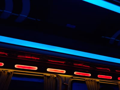 The image shows the interior of a night train. A row of red oblong lights illuminates the space above the window seats. Above this row is a blue LED strip light. Luggage racks are visible overhead in the dark space at the top of the image. Curtains partially cover the windows, and a hand appears near one of the windows. A small exit sign in a foreign language is visible on the ceiling. It is not possible to determine if this image depicts a scene within Anipemza, Armenia from the provided context.