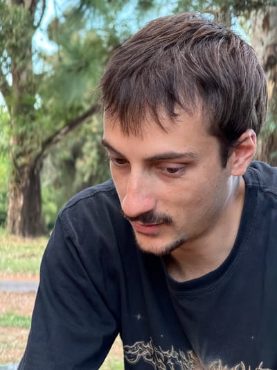 A young man with dark hair and a thin mustache is shown in a slightly angled portrait. He's wearing a dark-colored long-sleeved shirt with what appears to be a light-colored design near the collar. The background includes blurred greenery and a tree trunk, suggesting an outdoor setting. The lighting is soft, highlighting his features. His gaze is directed downwards, creating a pensive or contemplative mood.