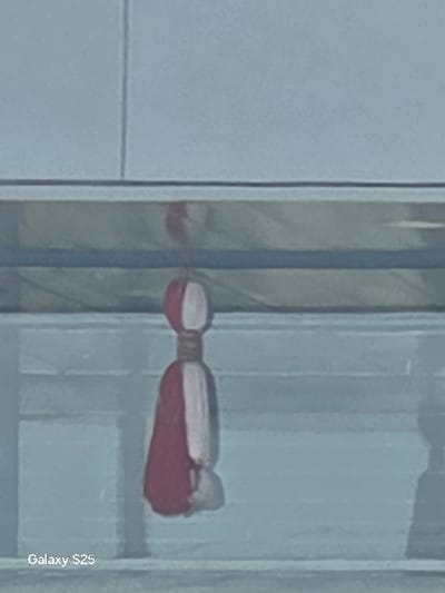 The image displays a single object, a red and white fabric tassel or charm, suspended vertically in the center of the frame. The object features a rounded top portion, bisected vertically into a white left half and a red right half. Below this, a narrower, bound section, likely dark cord or fabric, connects to a flared, skirt-like base composed of intermingled or striped red and white fabric segments. The object casts a faint shadow to its right.
The background is out of focus and light-toned, appearing to be an interior setting. A prominent horizontal element, likely a reflective surface such as a glass railing or partition, runs across the mid-ground behind the suspended object. This element shows a blurred, inverted reflection of the red and white object. Above this horizontal feature, the background is a plain light blue-grey. Below it, the background is also light blue-grey with subtle horizontal banding. No individuals are present. The bottom left corner of the image contains a watermark or metadata text: "Galaxy S25".
The specific location within the city of Novo Delchevo, Bulgaria, cannot be determined from the visual information provided in the image.