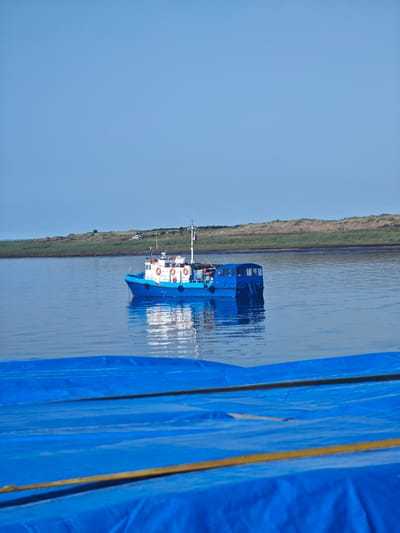 A medium-sized blue and white boat is anchored in calm, blue water. The boat has a white cabin with windows and a white upper deck. Several life preservers are visible on the side of the cabin. A Russian flag flies from a mast at the rear of the boat. In the background, a low, grassy, and sandy shore is visible, with a clear blue sky above. The foreground shows a large expanse of blue tarp, partially obscuring the view of the water and shore. This scene is likely in the vicinity of Dudinka, Russia, a port city located on the Yenisei River.