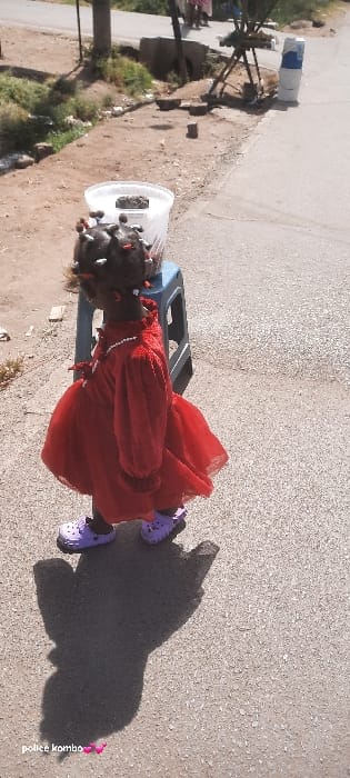 A child, viewed from behind, stands on a paved surface in Homa Bay, Kenya, under direct sunlight. The child wears a long-sleeved red dress with a tiered skirt, purple clogs, and a white beaded necklace. Their dark hair is styled in multiple small braids or twists, adorned with white and red beads. A pronounced shadow of the child is cast on the ground to their left. To the child's right and slightly behind, a blue plastic stool supports a translucent white plastic bucket with a dark, irregularly shaped object inside. The paved surface extends into the background, bordering a dirt area with sparse vegetation and visible debris. In the mid-ground, a dark vertical pole is visible. Further in the background, another paved or dirt road is visible, with multiple indistinct human figures and a makeshift wooden structure.
