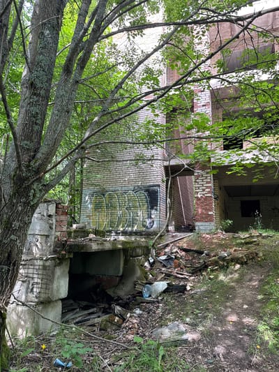 The image depicts an abandoned brick building overgrown with vegetation in a wooded area. The building appears to be multi-story with exposed concrete structures and damaged brickwork. Graffiti is visible on the exterior walls. The ground surrounding the building is littered with debris, including wood, bricks, and plastic bottles. The overall scene suggests a state of disrepair and neglect. The location is specified as Дубки Снт, Russia. There are no people visible in the image.