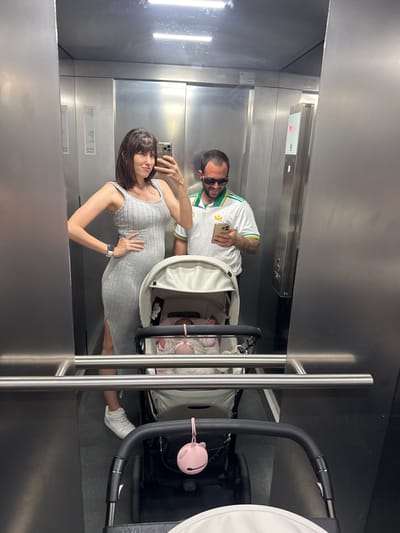 A couple in an elevator with a baby stroller are taking a selfie in a mirror. The woman is wearing a gray dress and white sneakers. The man is wearing a white soccer jersey with green and yellow accents and sunglasses. The stroller is white with a black frame. The elevator appears to be made of stainless steel. It is not possible to ascertain the location in Pomezia, Italy from the image.